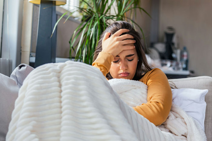 Young woman wrapped in a blanket sitting on a couch, looking upset and holding her forehead after a friendship of 10+ years ended.