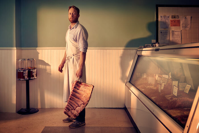 Butcher in an apron holding a large cut of meat in a shop, showcasing breathtaking food photography from the awards.