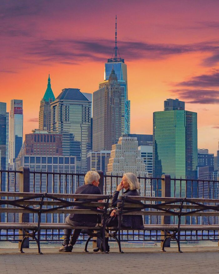 Couple sitting on park benches with New York City skyline during sunset in a creative photo edit making you question reality.