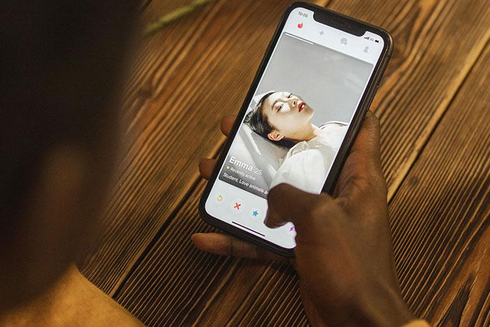 Person holding smartphone showing dating app profile with a woman’s photo and brief bio on wooden table background.