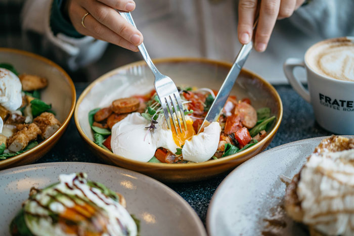 Person cutting into poached eggs at a bottomless mimosa brunch with plates of food and coffee on the table.