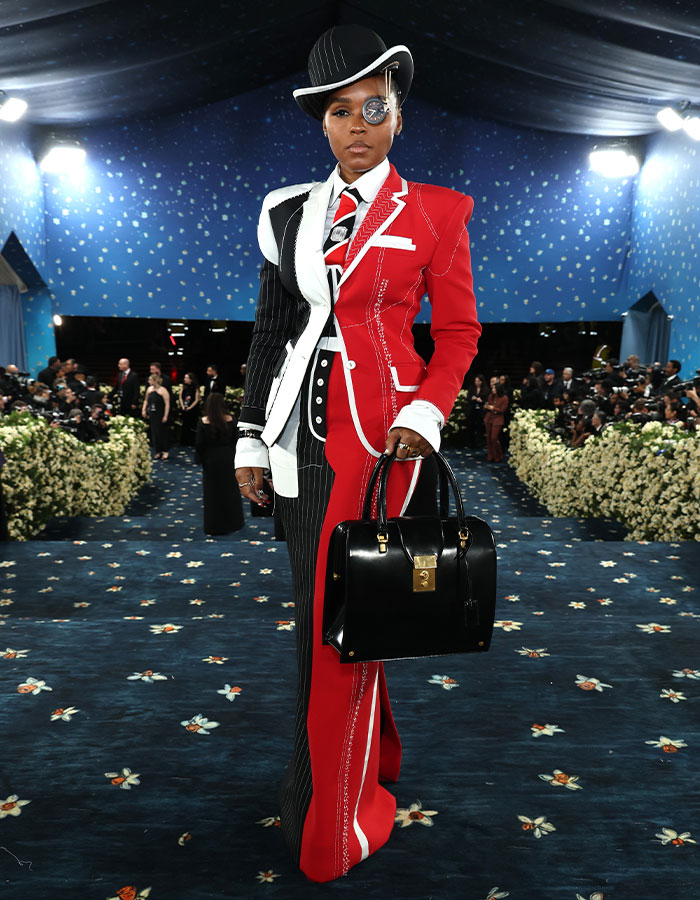 Person in a half-red, half-black pinstripe suit and hat with monocle, carrying a black handbag at a fashion event.
