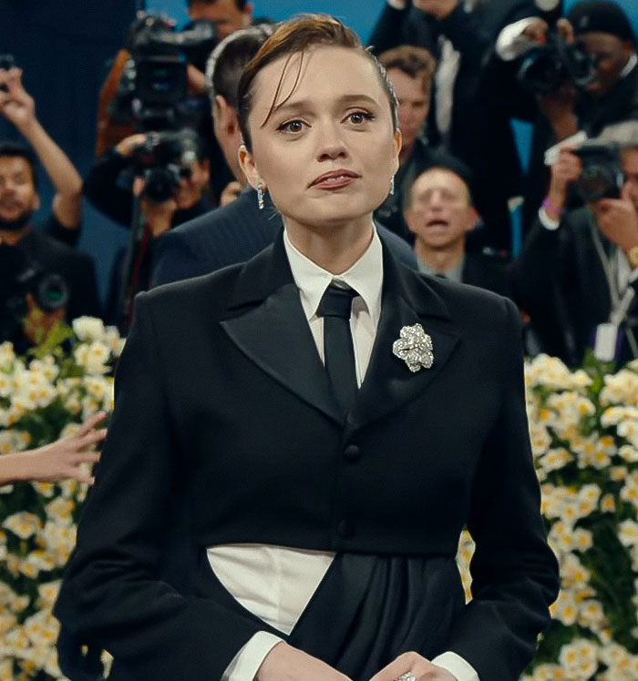 Aimee Lou Wood at an event, wearing a black suit with a brooch, attending a media gathering with photographers behind her. Aimee Lou Wood at an event, wearing a black suit with a brooch, attending a media gathering with photographers behind her.