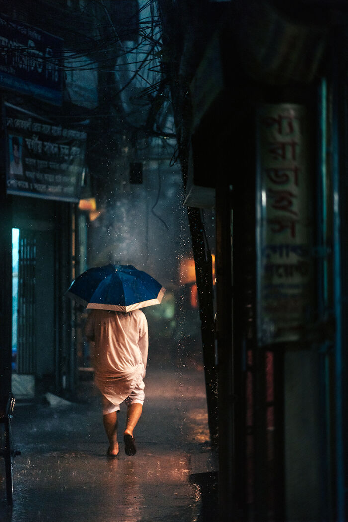Person walking barefoot in the rain holding a blue umbrella in a dimly lit narrow street street photography moment.