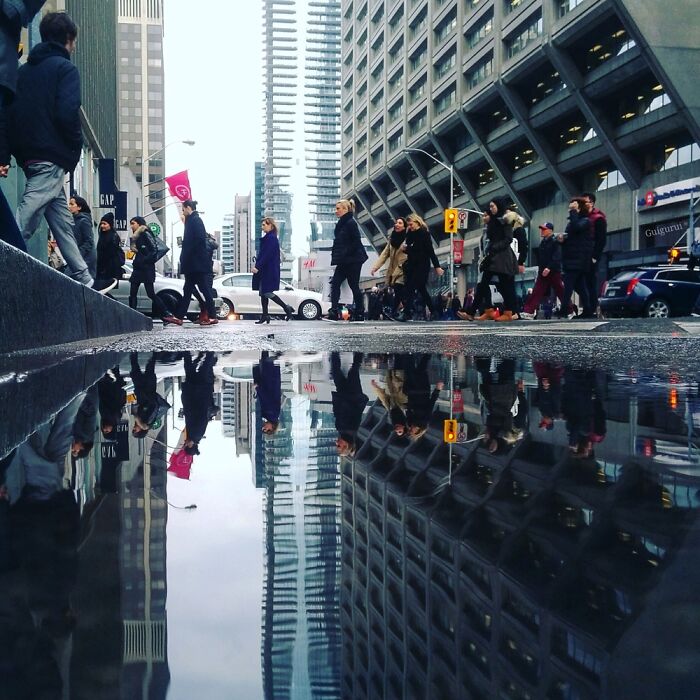 People crossing a city street with skyscraper reflections in a puddle, showcasing street photography moments by talented photographers.