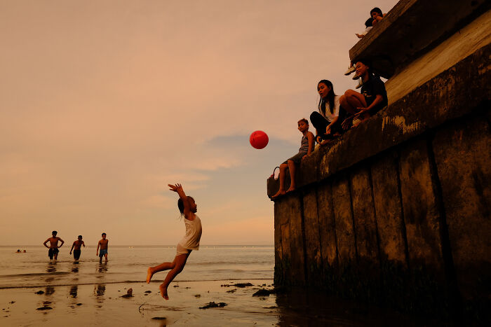 Child jumping to catch a red ball at sunset, with people sitting on a dock and others wading in the water, street photography moment.