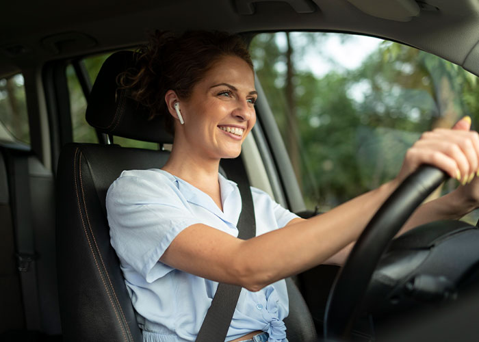 A woman wearing earbuds and smiling while driving a car, illustrating things everyone does but doesn’t talk about.