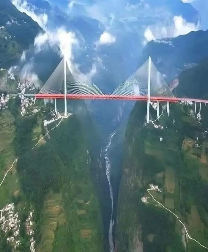 Aerial view of a long red cable-stayed bridge spanning a deep valley with green hills and clouds from Google Earth.