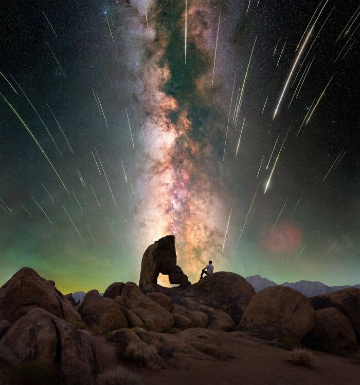 Person sitting on rocky landscape under mesmerizing Milky Way with shooting stars streaking across the night sky.