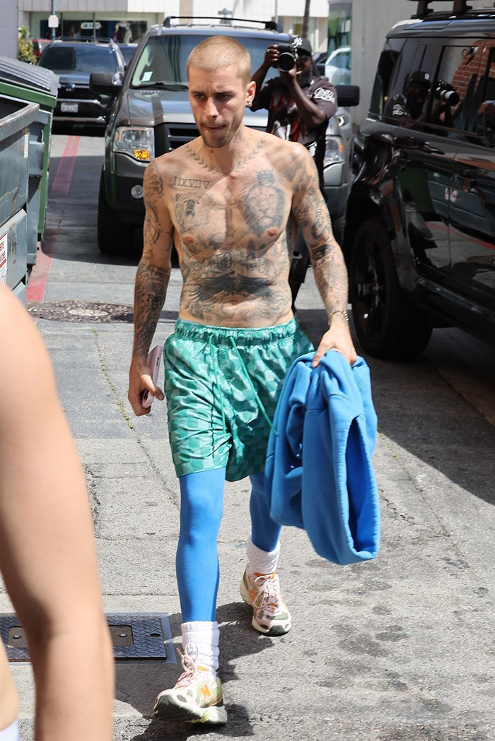 Justin Bieber shirtless walking in LA wearing green shorts and blue leggings while carrying a blue jacket outdoors.