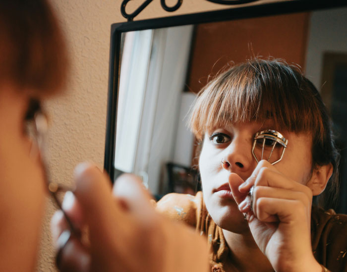 Woman using an eyelash curler in front of a mirror, illustrating problematic behaviors women get a pass for.