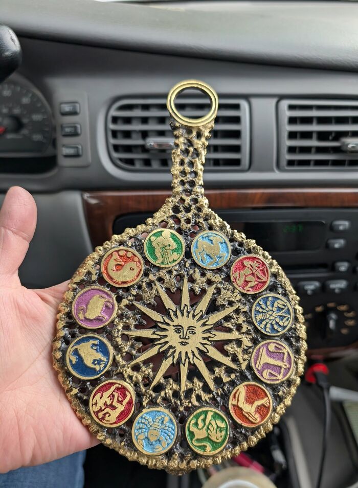 Vintage thrifted zodiac medallion with sun motif and colorful astrological symbols held inside a car interior.