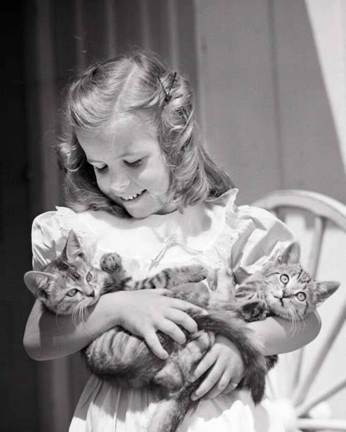Vintage black and white photo of a smiling girl holding two playful kittens, highlighting the timeless bond between kids and cats.