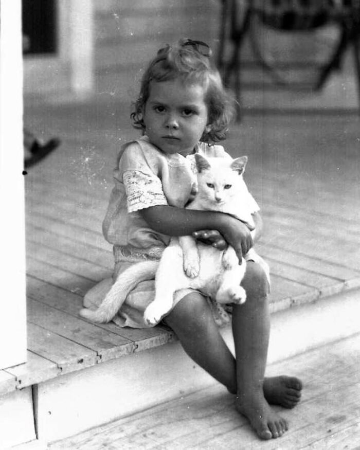 Vintage photo of a young child sitting on a porch, hugging a white cat, showcasing the timeless bond between kids and cats.