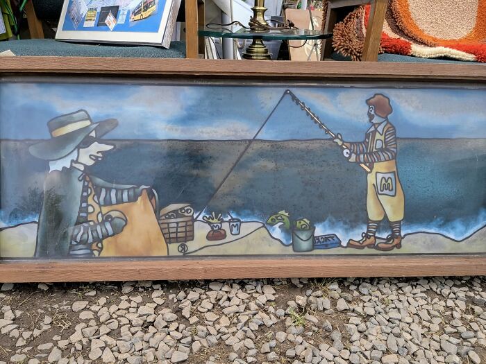 Framed thrifted art depicting a whimsical fishing scene with cartoon characters, a weird and wonderful find.