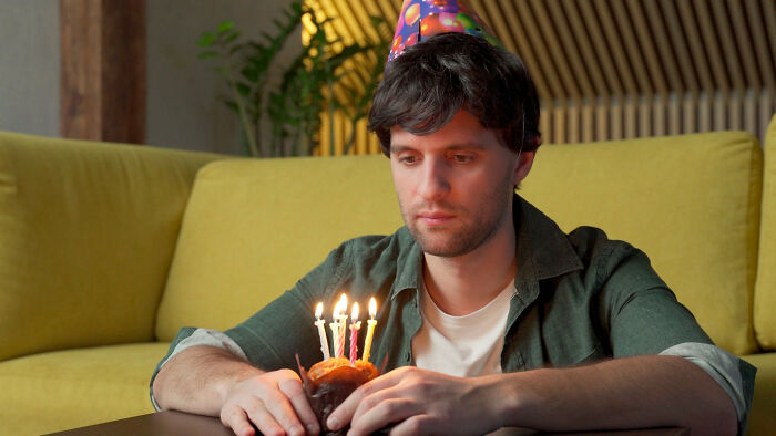 Young man wearing a party hat looks sad while sitting alone with a birthday cupcake, feeling like being picked last in gym class.