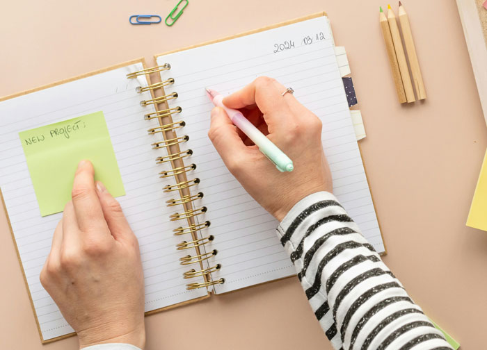 Person writing in spiral notebook with sticky note labeled New Project, illustrating bosses, teachers, or parents off the rails.