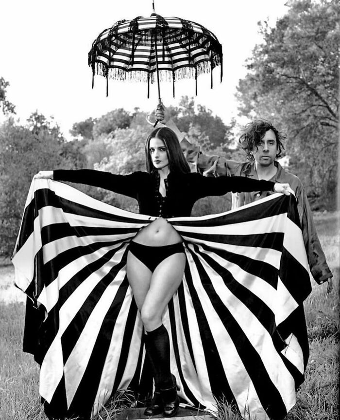 Black and white street photo featuring a woman with striped fabric and a man holding an umbrella in a natural setting.