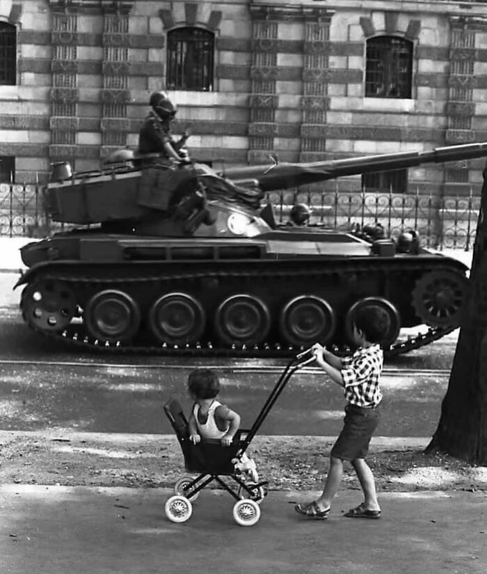 Black and white street photo showing two children playing with a stroller near a military tank on the street.