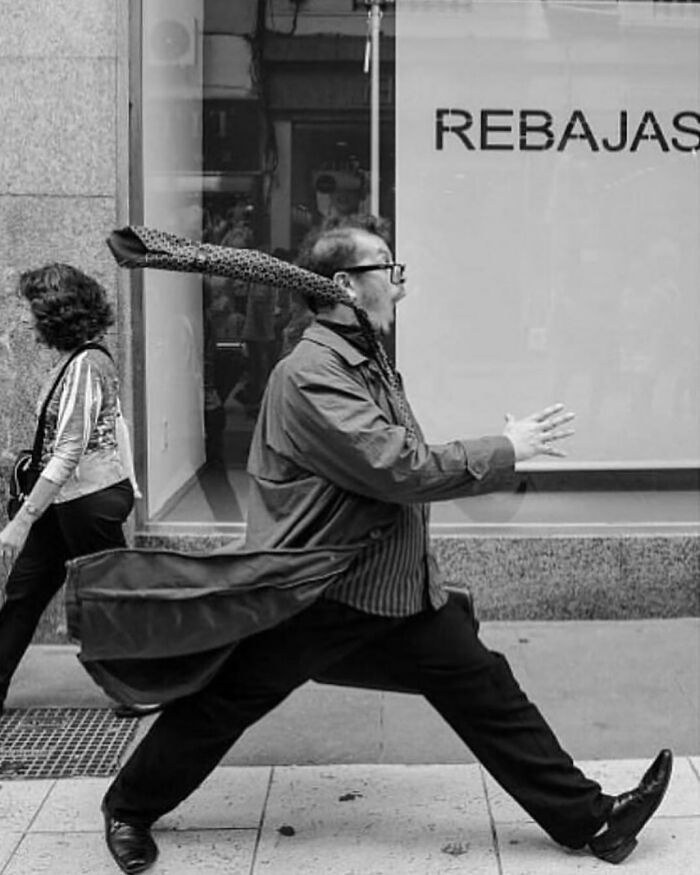Black and white street photo of a man walking briskly with a flowing coat and scarf on an urban sidewalk.