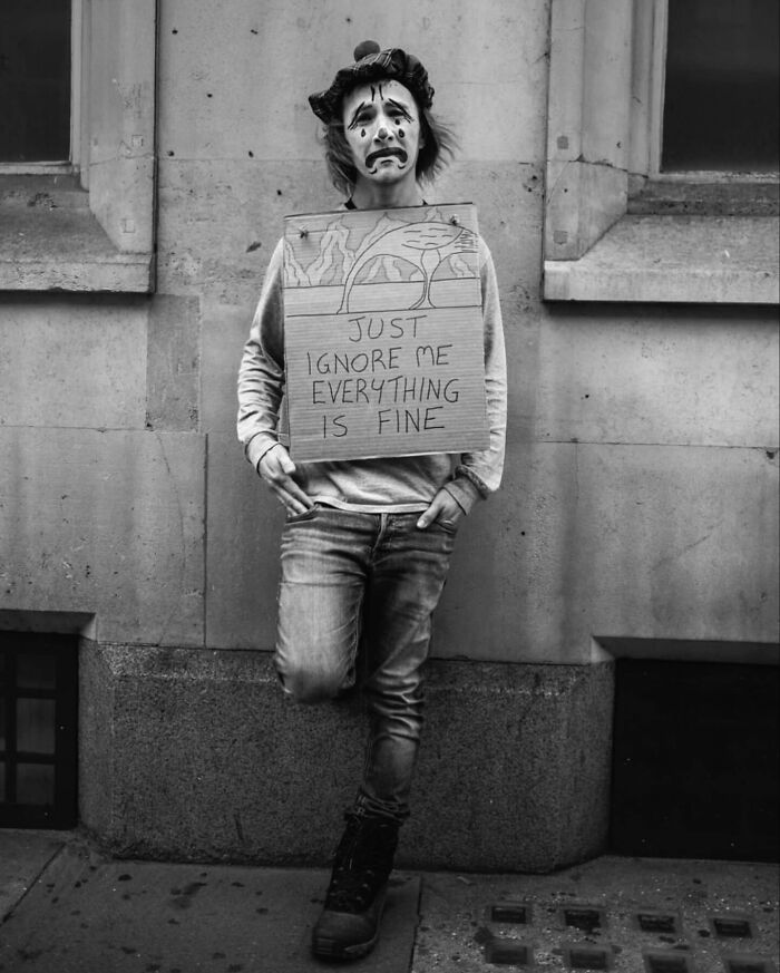 Black and white street photo of a person in clown makeup holding a sign with a message in an urban setting.
