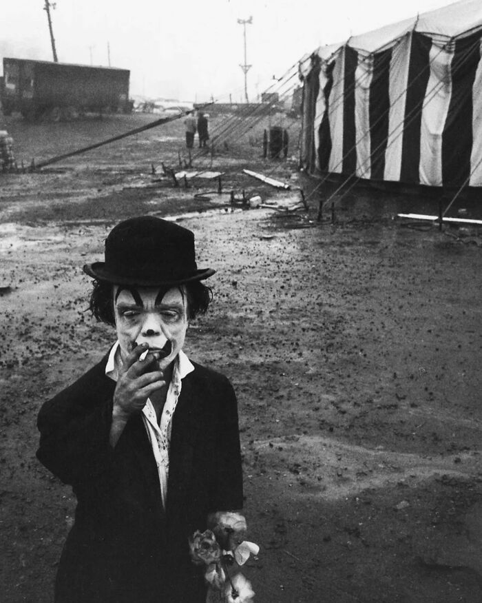 Black and white street photo of a clown smoking a cigarette near a striped circus tent on a muddy ground.