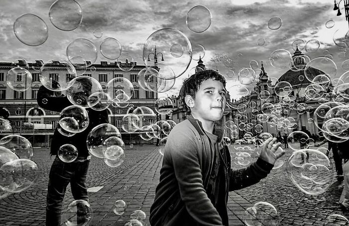 Black and white street photo of a boy surrounded by floating bubbles in an urban square with historic buildings.