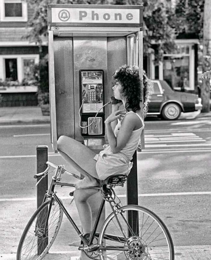 Black and white street photo of a woman sitting on a bicycle using a public phone in an urban setting.