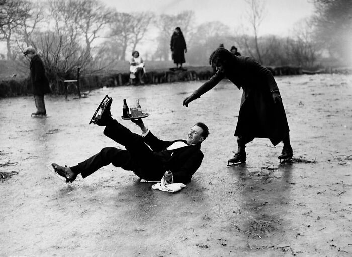 Black and white street photo showing a man slipping on ice while holding a tray and a woman reaching out to help.