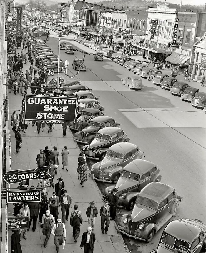 Black and white street photo showing vintage cars parked along a busy sidewalk with many pedestrians walking by.