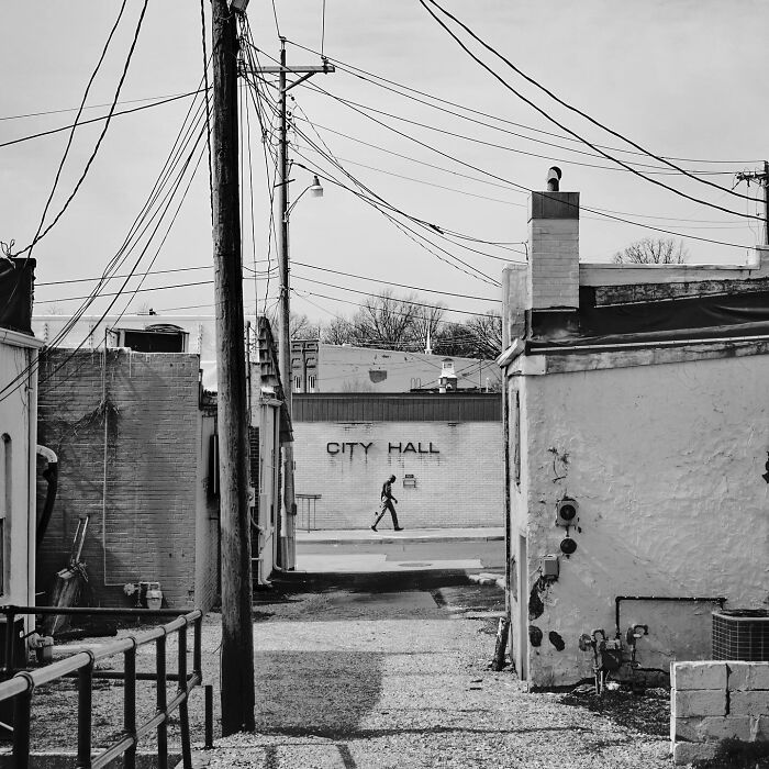 Black and white street photo showing an urban alley with power lines and a person walking near a city hall building.