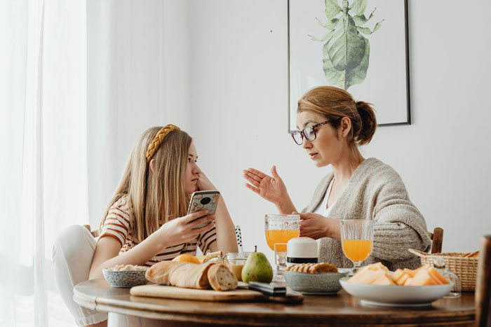 Woman in glasses explaining something to a girl with a phone at a breakfast table, illustrating infuriating things said by people in charge