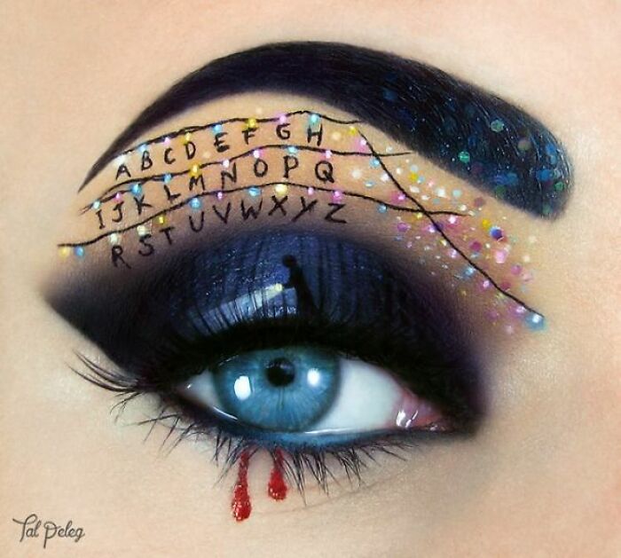 Creative makeup look featuring alphabet letters and string lights on eyelid with dramatic blue eyeshadow and detailed artistry.