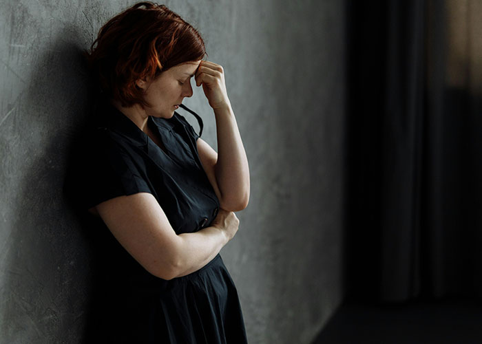 Woman with red hair in black dress standing against wall, looking distressed, reflecting trauma after newborn situation.