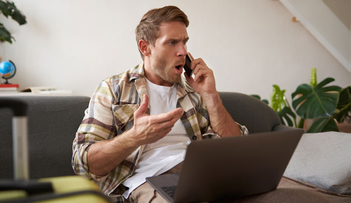 Man with a shocked expression talking on phone while using laptop, reacting to cousin offering wedding photography gift then billing $3,000