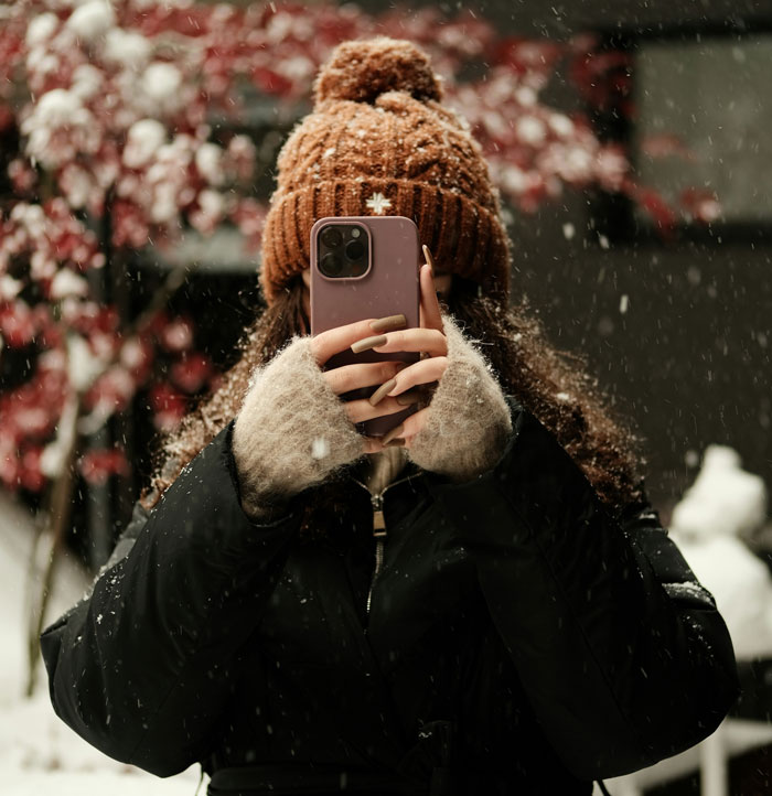 Woman in a winter coat and knit hat taking a photo with smartphone, illustrating problematic behaviors women get a pass for.