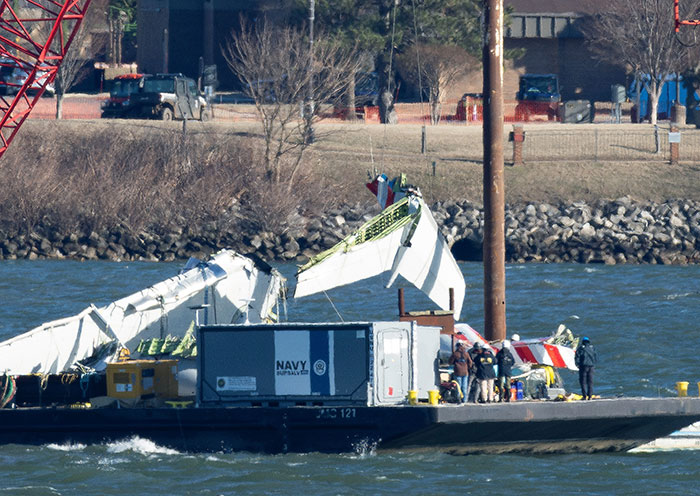 Wreckage of a DC plane crash on water with Navy salvage crew examining the debris near a shore with vehicles and trees.