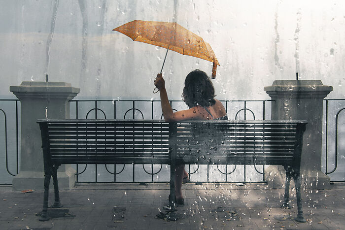 Person sitting on a bench holding an orange umbrella in rain, capturing a striking street photography moment.