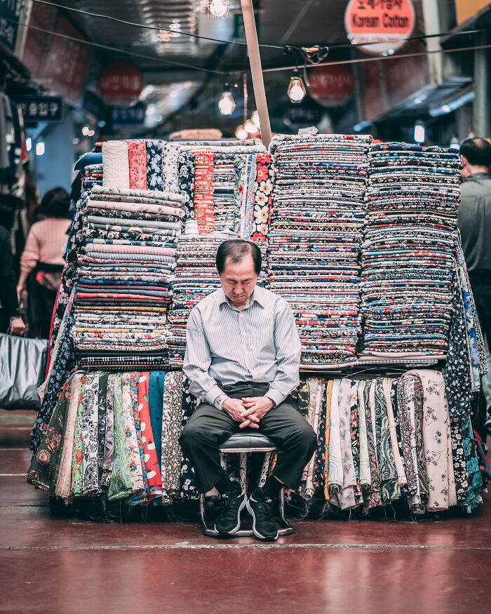 Man sitting in front of colorful fabrics at a market, capturing a street photography moment by talented photographers.