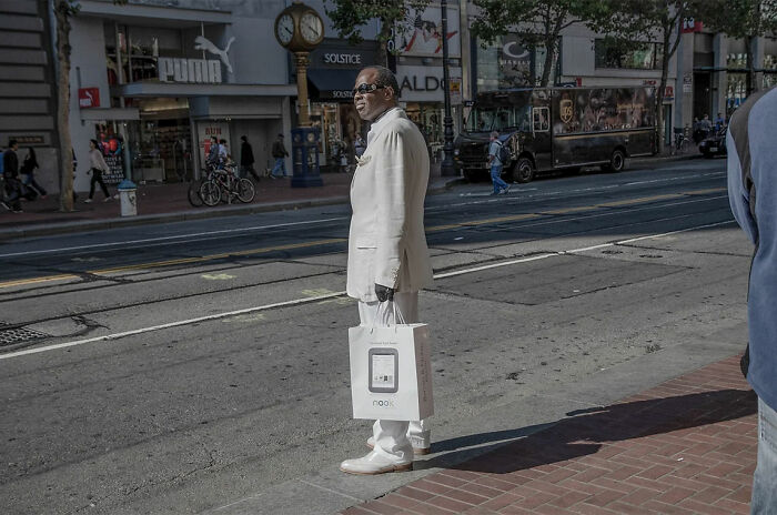 Man dressed in white standing on a city street holding a shopping bag in a street photography moment.