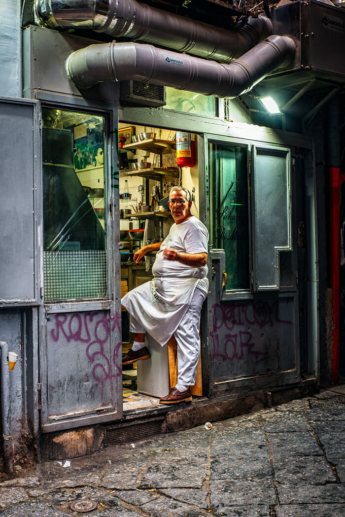 Street photography moment of a man in white apron standing in a doorway of a small urban kitchen with industrial pipes above.