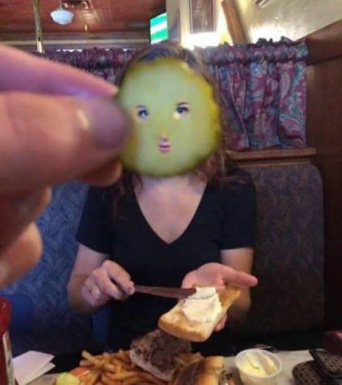 Hand holding a pickle slice with a funny face emoji covering a woman's face while she buttering bread at a restaurant.