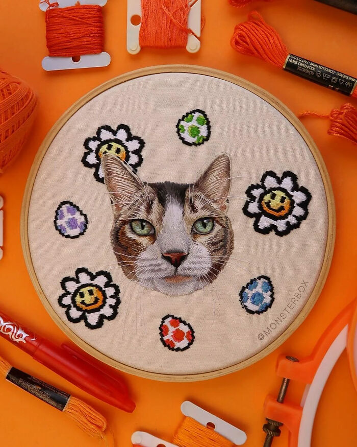 Realistic embroidery design of a cat's face surrounded by playful flowers and colorful egg patterns on fabric.