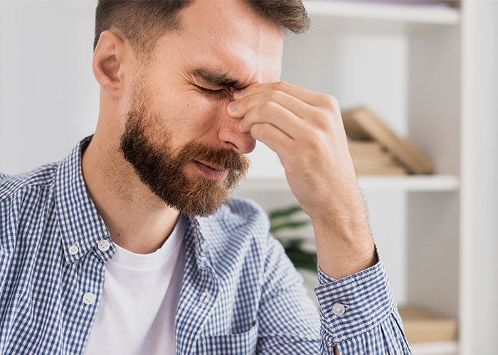 Man in a checked shirt pinching bridge of nose in discomfort, illustrating the most pathetic injuries shared by people.