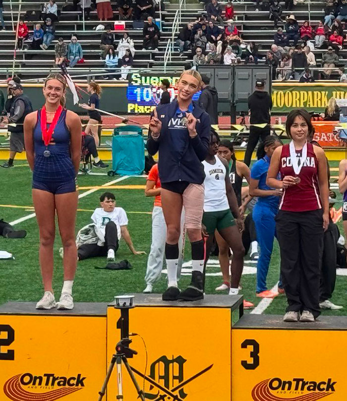 High school girl stands defiantly on first-place podium at track event with medals and athletes in the background