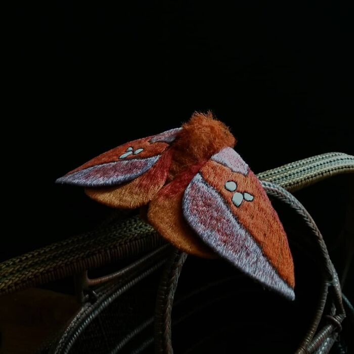 Embroidered fiber moth with detailed orange and gray wings, showcasing intricate textile art on a dark background.