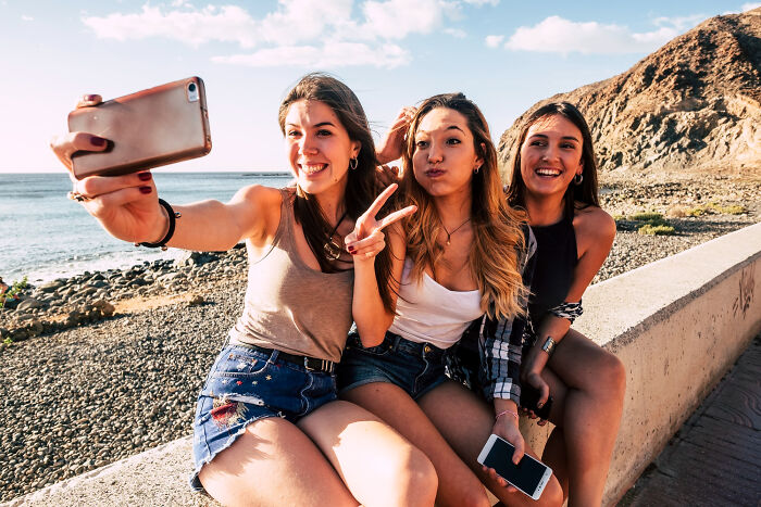 Three young women sitting by the beach taking a selfie, capturing moments that feel like being picked last in gym class.