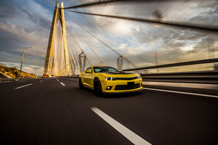 Yellow sports car driving on a modern bridge under a dramatic cloudy sky during sunset.