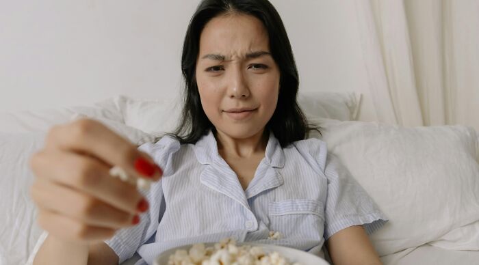 Young woman in pajamas on sofa holding popcorn, focusing intently, testing her Hollywood movie knowledge quiz.