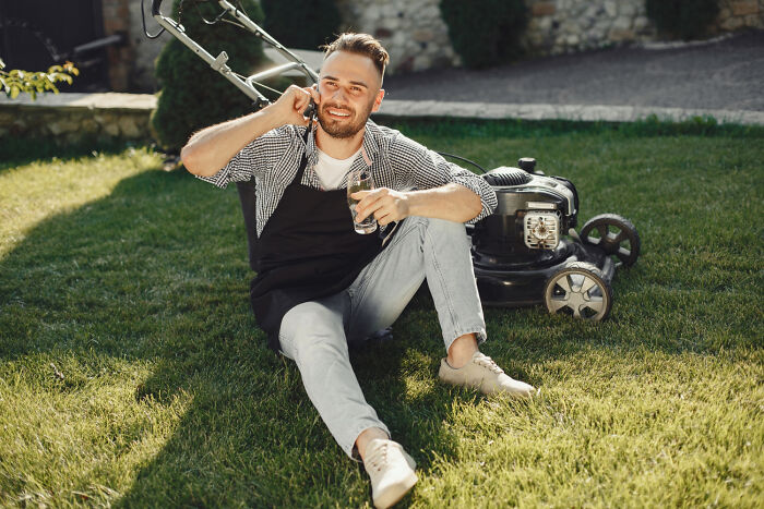 A man sitting on grass near a lawn mower, holding a glass and talking on the phone in a sunny backyard.